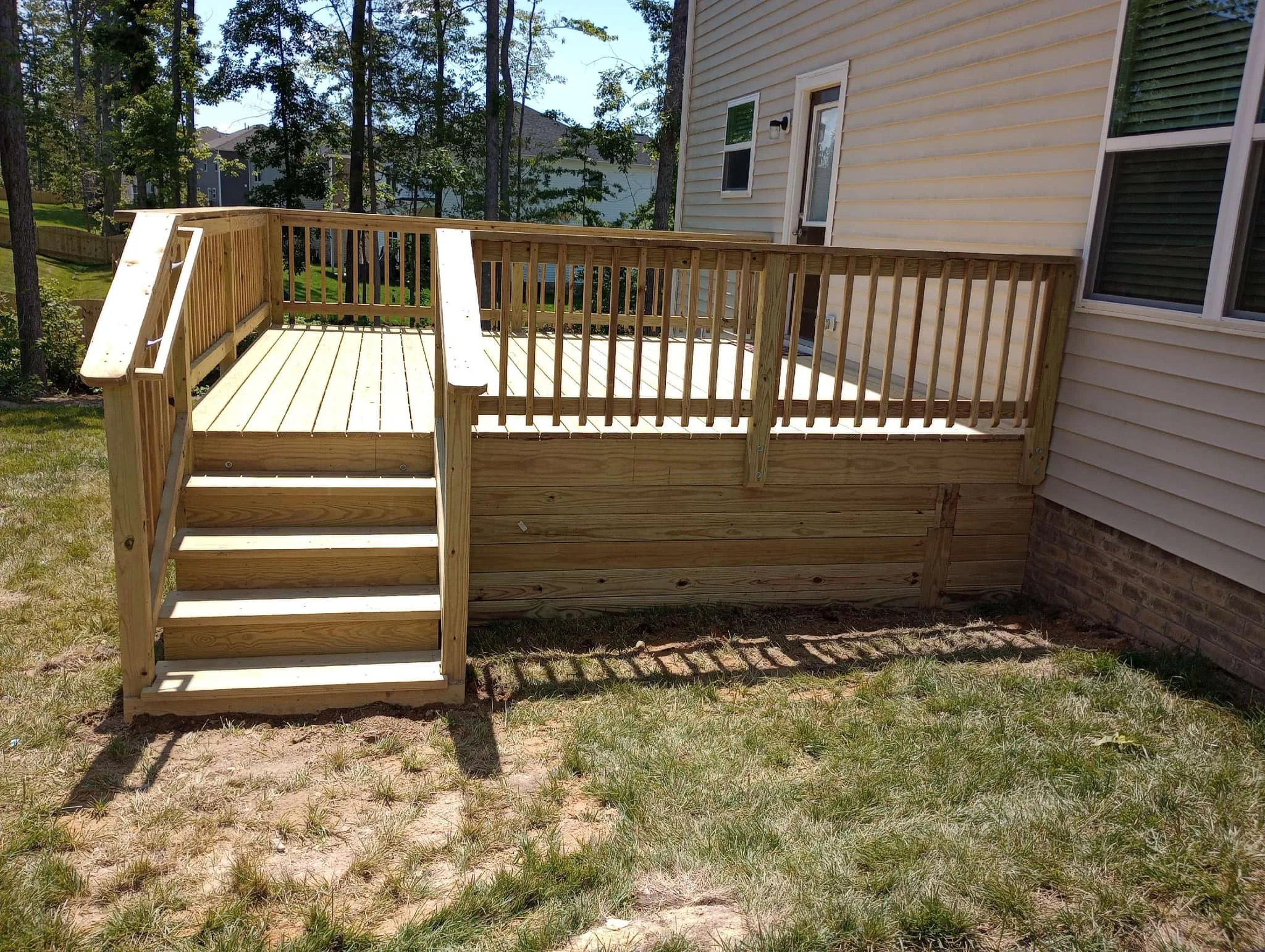 A newly built wooden deck attached to a beige house, featuring clean, uniform boards and a sturdy railing, illustrating a full deck replacement rather than resurfacing.